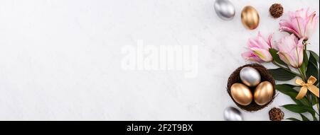 Golden and silver Easter eggs in the nest with pink lily flower on bright marble white table background. Stock Photo
