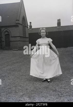 1956, historical, a young girl in the grounds of a church showing off her frock or gown she will wear for the traditional May Day parade or carnival, Leeds, England, UK. An ancient celebration of springtime, May Day is believed to date back to Roman times and Floralia, the festival of Flora, the Roman goddess of flowers. Modern May Day traditions, in both Europe and USA, include dancing around the maypole and the crowning of the Queen of May after a parade. Stock Photo