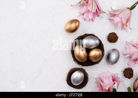 Golden and silver Easter eggs in the nest with pink lily flower on bright marble white table background. Stock Photo