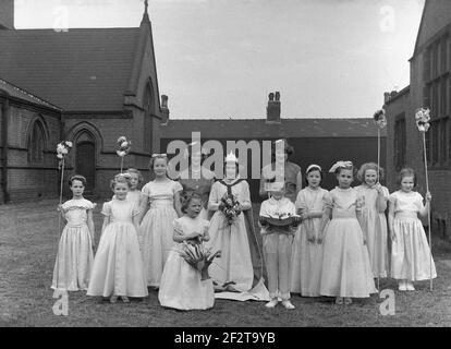 1956, historical, in the grounds of their local church, a group of children in their pretty frocks or dresses, line-up for a photo before the traditional May Day parade or carnival, Leeds, England, UK. The May Queen stands in the middle of the group, with a little boy holding a cushion, which held her crown. Several of the girls are holding flower sticks. The May Day celebrations are a traditional festival, marking the beginning of summer, dating back to Roman times, and the festival of Flora. Stock Photo