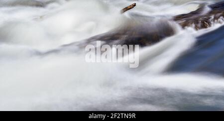 Long exposure photo - water flowing over rock near drifted tree branch, everything smooth, only stationary wood and rock is in focus Stock Photo