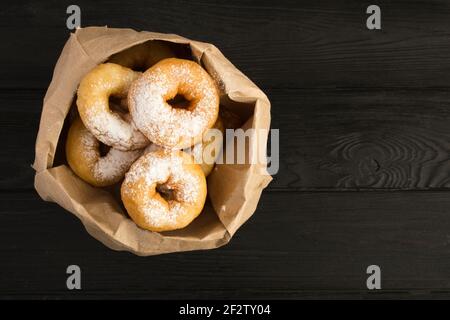 Homemade donuts with powdered sugar in the paper package  on the black wooden background. Top view. Copy space. Stock Photo
