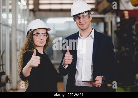 Portrait of Two Machinists is standing with confident with   safety helmet in front the glass wall of industry factory. Stock Photo
