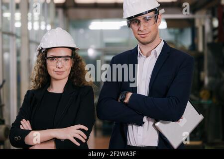 Portrait of Two Machinists is standing with confident with   safety helmet in front the glass wall of industry factory. Stock Photo