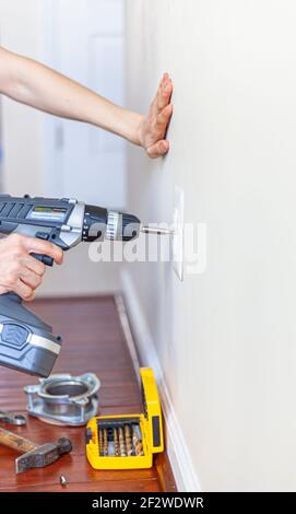 A woman is using a cordless electrical drill with screwdriver bit attached to remove the screw holding the cover plate of an electrical outlet on the Stock Photo