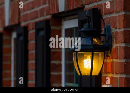 A coiled compact fluorescent light bulb glowing in dark. The bulb is inside a metal wall lantern attached to the brick wall of the exterior of a house Stock Photo