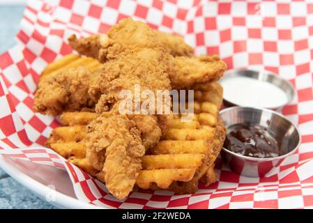 Chicken tenders breaded and cooked to perfection sitting on top of a pile of waffle french fries along with dipping sauces. Stock Photo