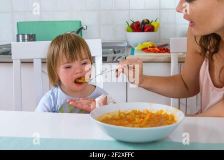 Mom feeding her disabled child with down syndrome with a spoon. Boy doesn't like offered food. Mother giving food to toddler at home. Baby food. Stock Photo