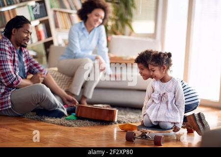 Kids riding skateboard on the floor and enjoying the playtime with their parents in a cheerful atmosphere at home. Family, together, love, playtime Stock Photo