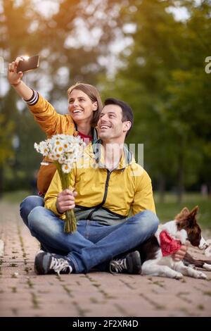 young caucasian woman taking selfie of herself, her boyfriend holding bucket of flowers, and their dog, outdoor in a park Stock Photo