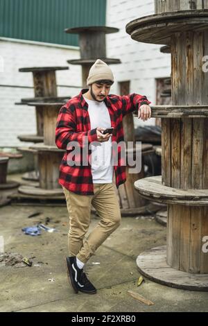 A vertical shot of a stylish Spanish male using his phone on background of industrial spools Stock Photo