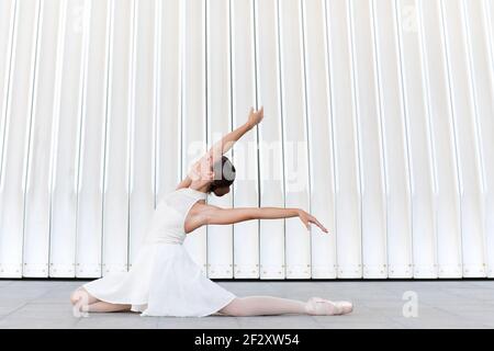 Side view of young graceful ballerina in white dress sitting on walkway while performing ballet pose and looking up Stock Photo