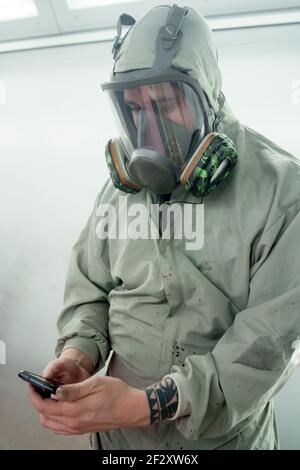 Male worker in respirator and protective uniform standing in car service and browsing mobile phone Stock Photo