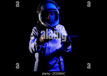 Male cosmonaut wearing white space suit and helmet while standing on black background in blue neon light holding small satellite Stock Photo