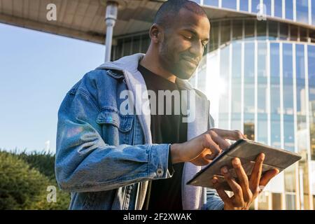Crop content adult African American male browsing internet on tablet against contemporary building in town in sunlight Stock Photo
