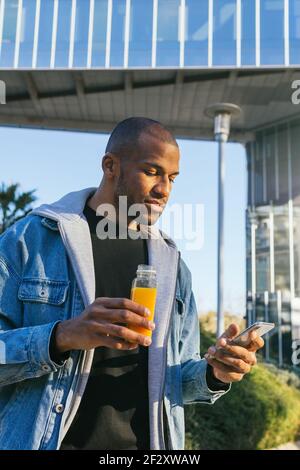 Adult ethnic African American male with bottle of orange juice surfing internet on cellphone in city Stock Photo