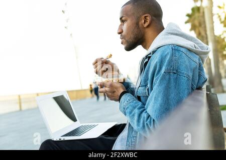 Side view of crop dreamy ethnic male distance worker with delicious lunch in container sitting with netbook in city and looking away Stock Photo