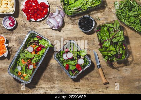 Top view composition of delicious vegetable salads in foil bowls placed on table near various ingredients including cherry tomatoes onions radish and Stock Photo