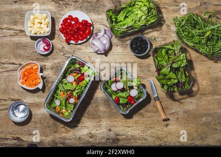 Top view composition of delicious vegetable salads in foil bowls placed on table near various ingredients including cherry tomatoes onions radish and Stock Photo