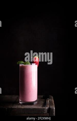 Delicious healthy homemade milkshake with fresh strawberry garnished with green mint leaves served in glass on wooden table against black background