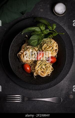 Top view of palatable Italian tagliatelle pasta with pesto sauce garnished with fresh red tomatoes and basil leaves served on black plate Stock Photo