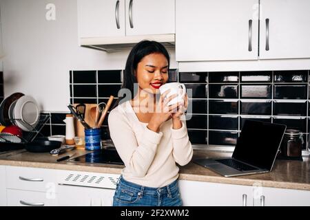 Smiling young Hispanic lady wearing casual outfit standing near kitchen counter with portable laptop with eyes closed holding ceramic cup Stock Photo