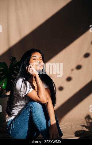 Attractive young Hispanic Woman wearing white cloth touching face with eyes closed in sun shadow Stock Photo