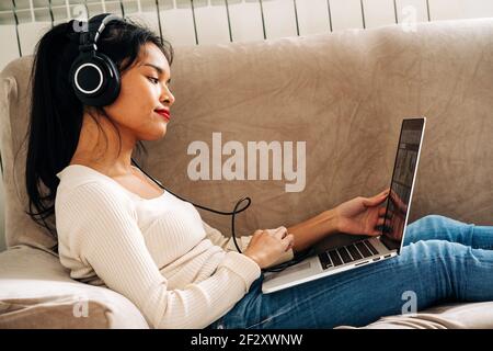 Side view of focused ethnic female lying on sofa with headphones working on modern laptop Stock Photo