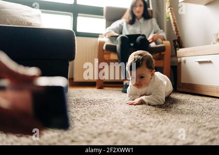 Cute baby crawling on carpet towards parent showing funny video on mobile phone while spending time together in living room Stock Photo