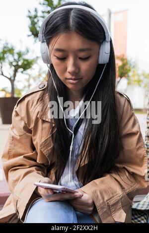 Young focused Asian female in casual clothes sitting on street with headphones browsing internet on mobile phone Stock Photo