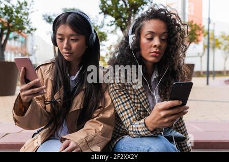 Pensive Asian female having fun call on smartphone and focused black Woman listening to music and surfing internet on mobile phone Stock Photo