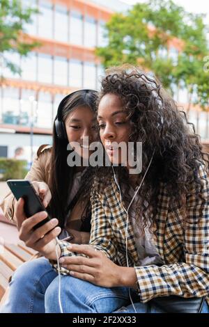 Asian female having fun call on smartphone and focused black Woman listening to music and surfing internet on mobile phone Stock Photo