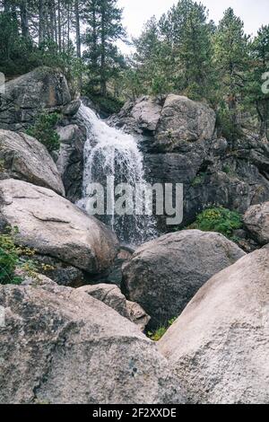 Waterfall of mountain river flowing over big stones between trees on cliffs in gorge Stock Photo
