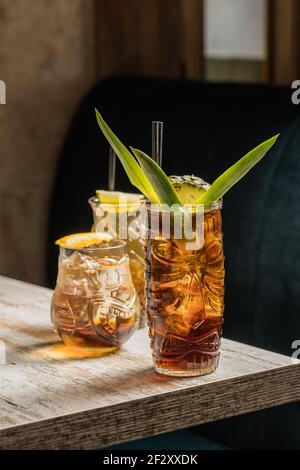 Glasses tiki mug filled with alcohol beverage with straw decorated with fruit placed on edge of wooden table couch Stock Photo