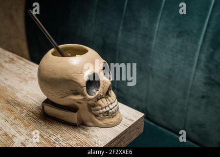 Ceramic polynesian tiki cup skull shaped with straw placed on wooden table on blurred background Stock Photo