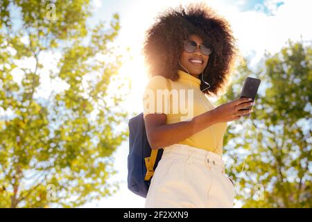 From below of glad ethnic female in earphones and sunglasses taking selfie on cellphone under blue sky in sunlight Stock Photo
