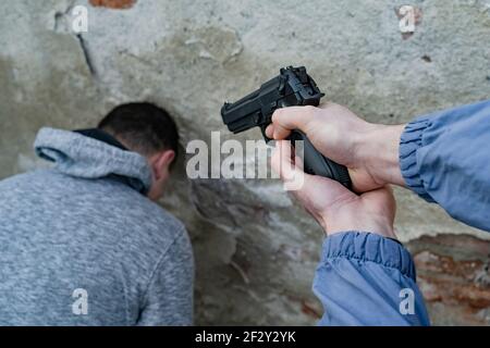 Close up on hand of unknown man terrorist or murderer holding a gun pointed to the head of unknown victim hostage with head leaned against a wall - te Stock Photo