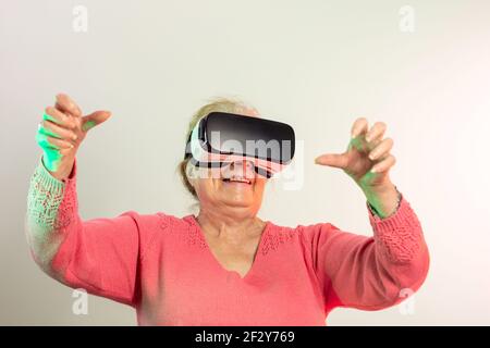 smiling senior woman in pink sweater with virtual reality glasses and raised hands looking straight ahead illuminated with red and green lights on lig Stock Photo