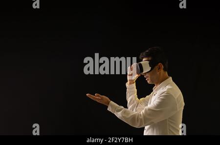 young guy in white shirt and virtual reality glasses surprised looks at his hand on black background Stock Photo