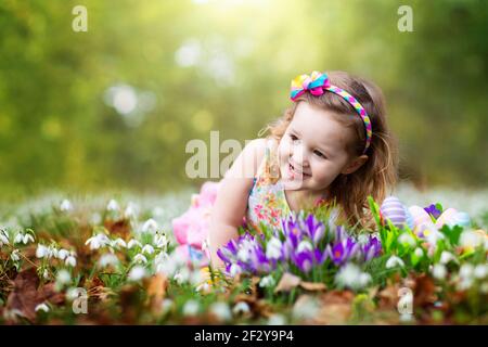 Child on Easter egg hunt in blooming garden with spring flowers. Kid with colored eggs in basket. Little girl picking flower. Stock Photo