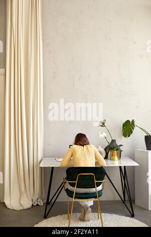 Back view of unrecognizable female in smart casual clothes sitting at desk and browsing data on netbook with empty screen while working Stock Photo