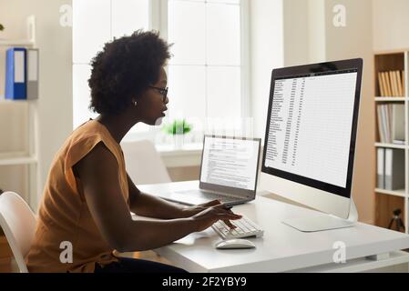 Young African-American woman sitting at office desk and working with data on computer Stock Photo