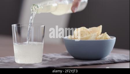 man pour ginger beer into tumbler glass with blue bowl full of potato chips, wide photo Stock Photo