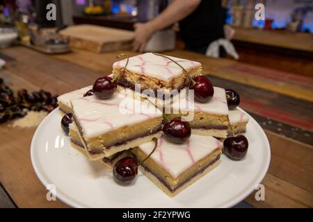A delicious plate of Cherry Bakewell Squares Tarts on a wooden kitchen table surrounded by cherries Stock Photo
