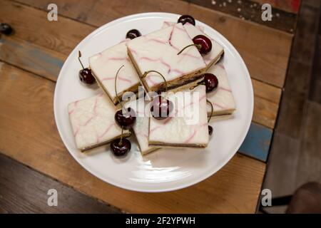 A delicious plate of Cherry Bakewell Squares Tarts on a wooden kitchen table surrounded by cherries Stock Photo