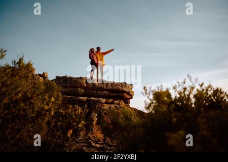 Young man standing at cliff peak pointing and showing something to woman during sunrise Stock Photo