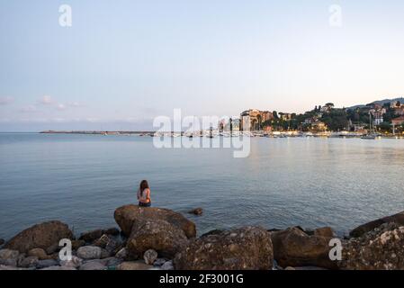 Young girl is sitting on he rocks and watching Rapallo harbour by night. Summer evening in Rapallo, Liguria, Italy Stock Photo