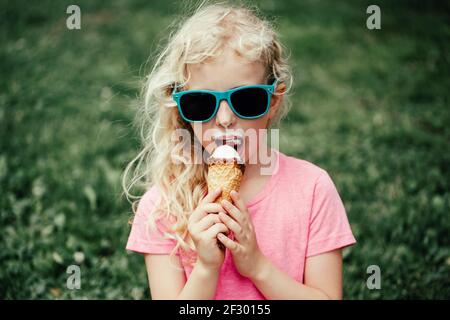 Cute funny adorable girl in sunglasses with dirty nose and moustaches eating ice cream from waffle cone. Happy cool child eating licking tasty sweet Stock Photo