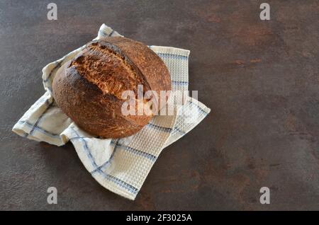 Freshly baked loaf of rye artisan bread on a kitchen towel on a dark background close-up. Stock Photo