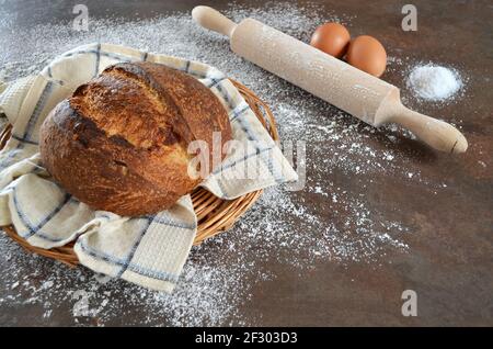 Freshly baked homemade sourdough bread with a crisp crust on a kitchen towel on the kitchen table sprinkled with flour. A rolling pin, two eggs, and a Stock Photo